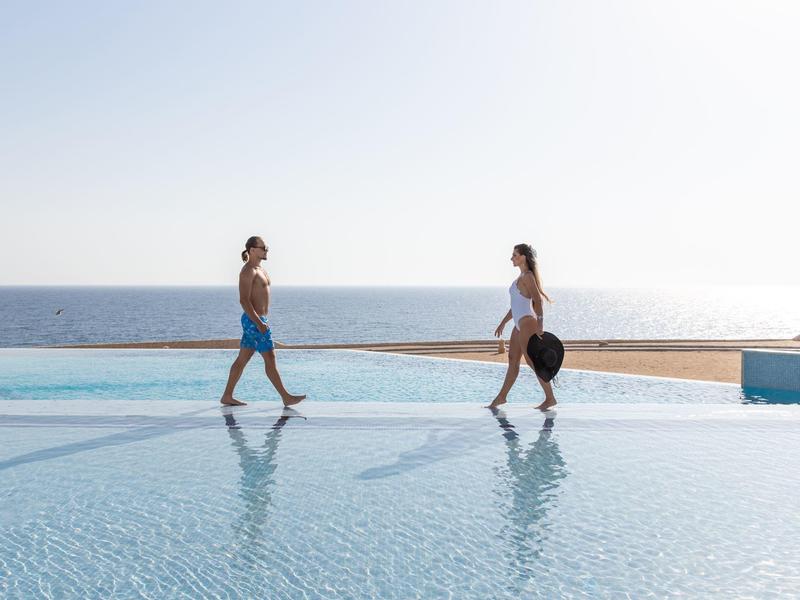 Couple walking by an infinity pool overlooking the calm ocean on a sunny day.