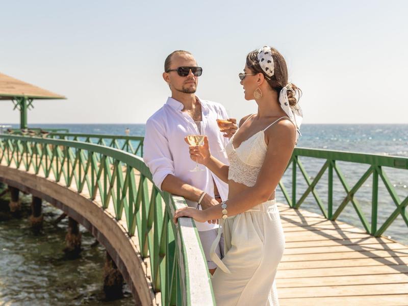 Couple enjoys drinks on a sunny boardwalk by the sea with green railings and clear sky.