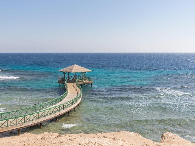 Curved wooden pier with a thatched roof gazebo over clear blue sea and sandy shore