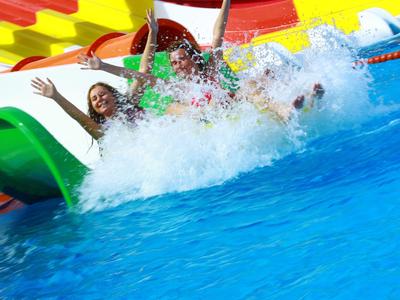 Drei Personen rutschen lachend eine bunte Wasserrutsche ins klare blaue Wasser.