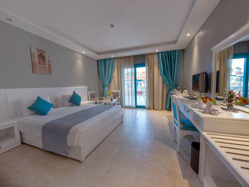 Helles Hotelzimmer mit Doppelbett, blauen Kissen, Schreibtisch, Fernseher und Balkon mit Vorhängen.