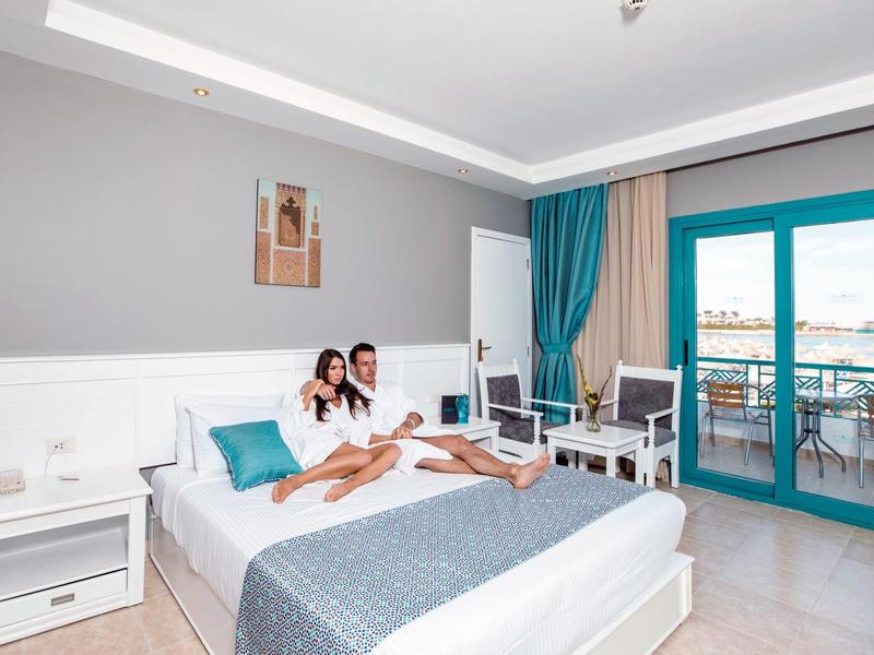 Helles Schlafzimmer mit grauen Wänden, weißem Bett, blauen Vorhängen und Balkon mit Meerblick.