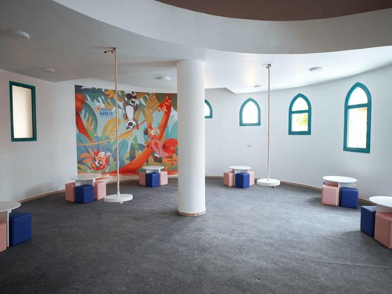 Großer Raum mit grauem Boden, runden Säulen, farbigen Sitzwürfeln und buntem Wandbild.