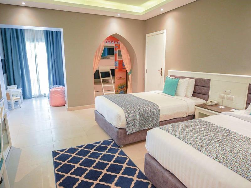 Helles Hotelzimmer mit zwei Betten, blauen Akzenten, Teppich und Balkon mit Vorhängen.