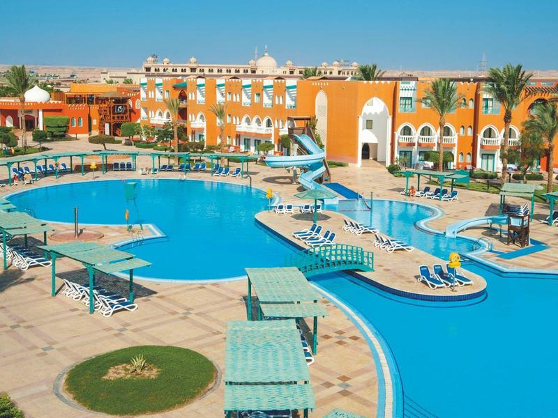 Großer blauer Pool mit Sonnenschirmen und orangefarbenem Hotel im Hintergrund an sonnigem Tag.