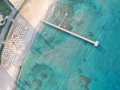 Luftaufnahme eines langen Stegs, der ins klare türkisfarbene Wasser und an einen Strand mit Liegestühlen führt.