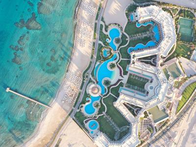Luftaufnahme von Hotel mit geschwungenen Pools, Strand, klarem türkisfarbenem Wasser und Grünflächen.
