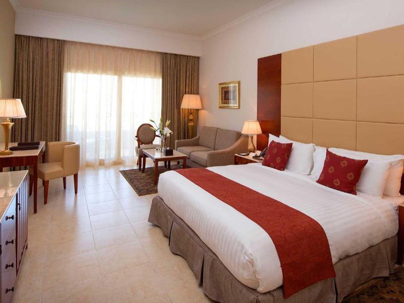 Habitación de hotel luminosa con cama doble grande, acentos en rojo y blanco y vista al balcón con sillas.