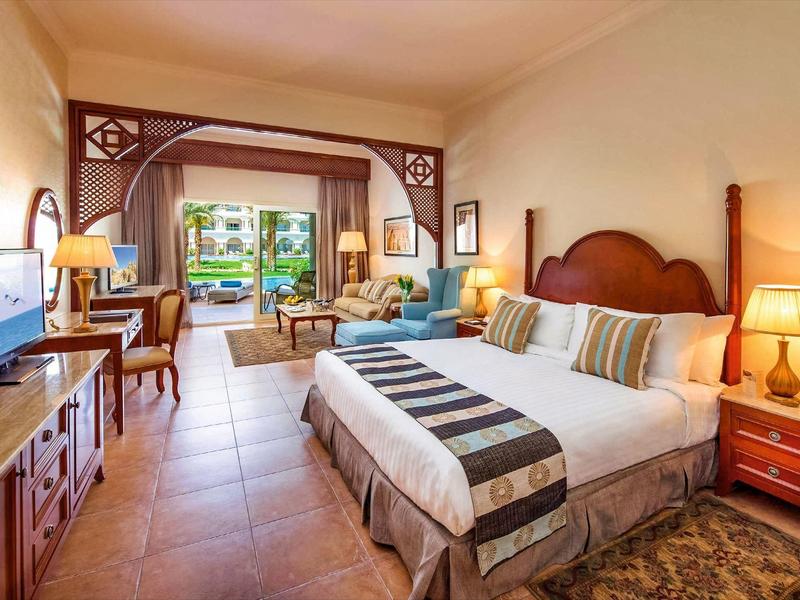 Habitación de hotel luminosa con cama doble, área de descanso y vista a terraza y jardín.