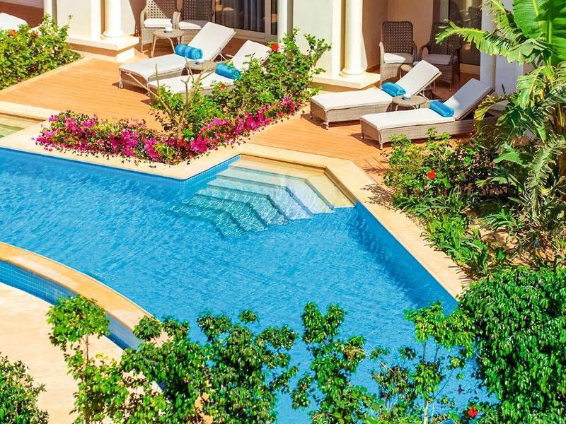 Piscina azul con jardín y tumbonas en terraza soleada de hotel con arcos blancos.