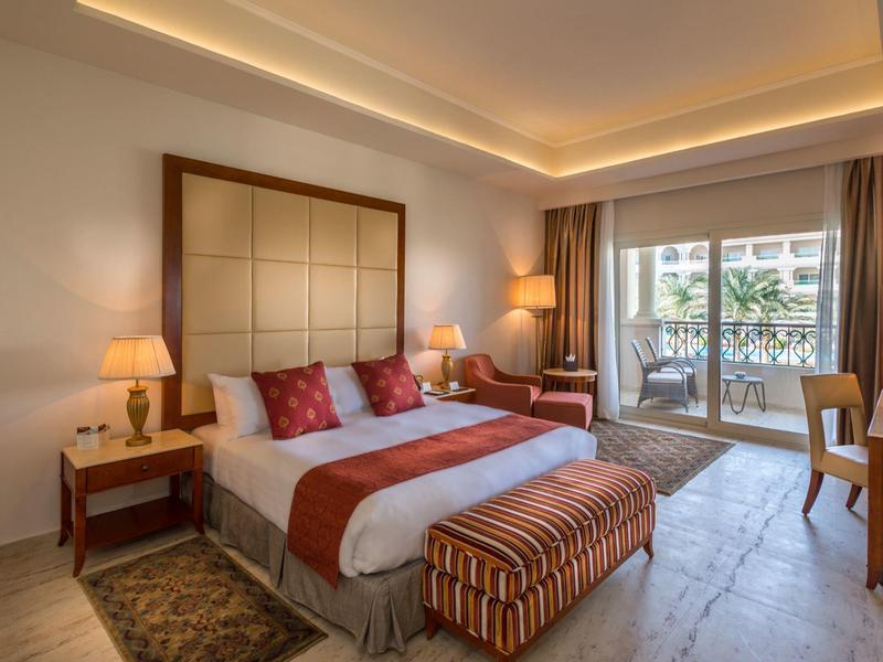 Habitación de hotel luminosa con cama doble, sillones, escritorio y balcón con vista al exterior.