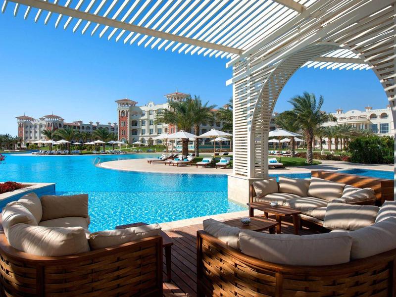 Lujosa terraza junto a la piscina con sillones giratorios, pérgola cubierta y vistas a edificios y palmeras del hotel.