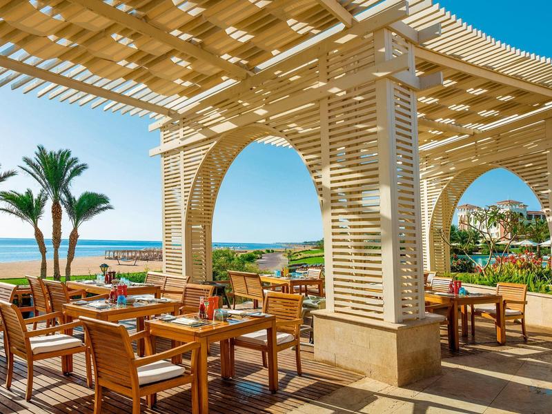 Área exterior abierta de un restaurante con muebles de madera y vistas al mar y palmeras bajo una pérgola.