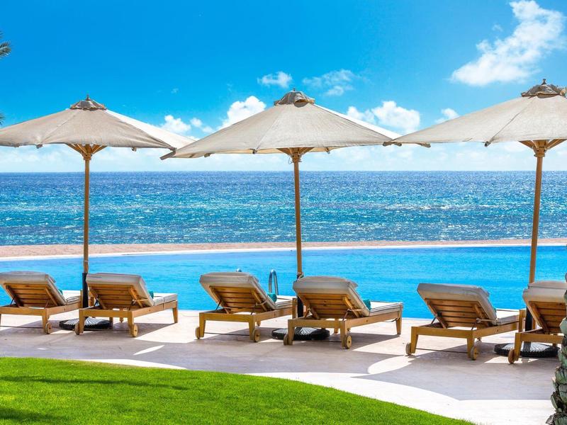 Área de piscina con tumbonas y sombrillas junto al mar bajo cielo azul.