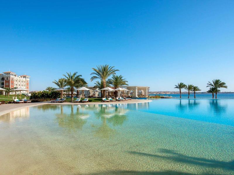 Piscina infinita con agua clara, palmeras y edificios de hotel bajo un cielo soleado.