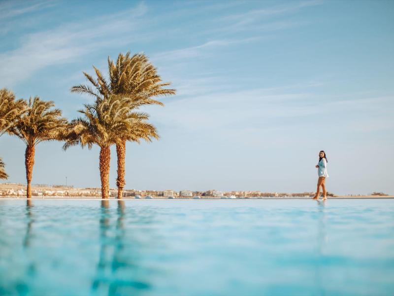 Mujer al borde de una piscina con palmeras y cielo azul de fondo.