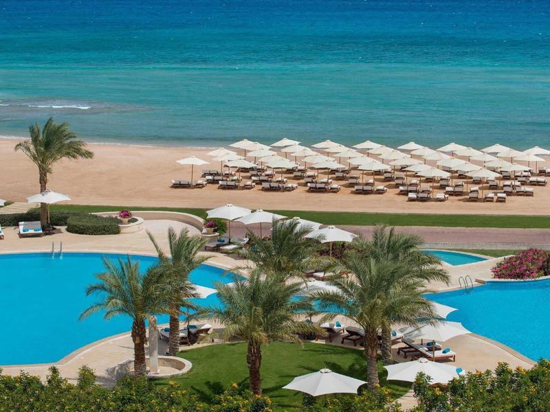 Piscina azul con palmeras y tumbonas junto a playa de arena y mar claro.