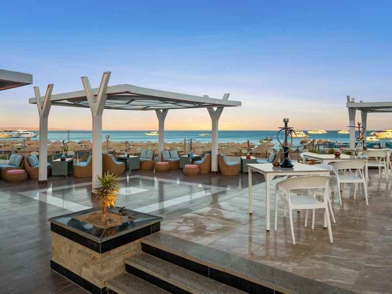 Dachterrasse mit Sitzbereichen, weißen Tischen, Holzpergolen und Blick auf das Meer bei Sonnenuntergang.