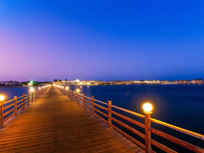 Long wooden pier with railings and lights extends over calm water at dusk.