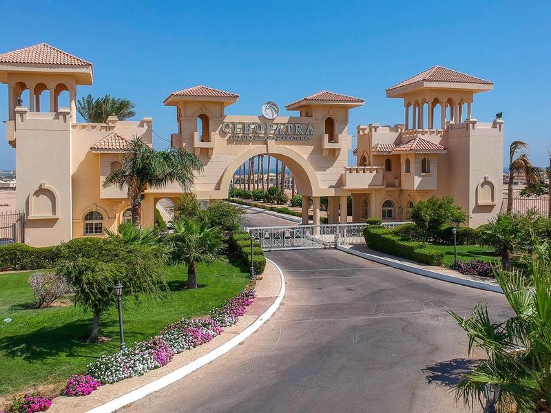 Entrance to a luxurious resort with arched gates, towers, palm trees, and landscaped gardens.