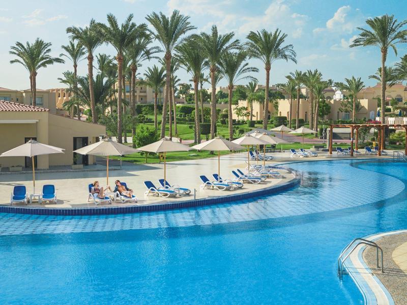 Large blue pool with lounge chairs and palm trees in a resort under a blue sky.