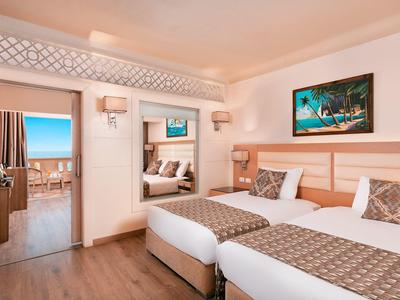Bright hotel room with two twin beds, wooden flooring, and a balcony view of the sea.