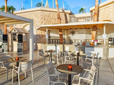 Moderní venkovní bar s stoly a židlemi vedle bazénu za slunečného počasí.