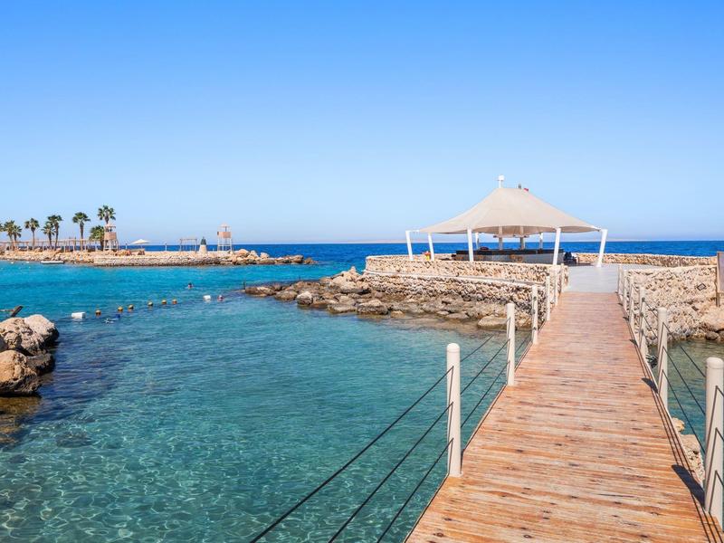 Wooden pier leading to a beach pavilion with clear blue water and palm trees in the background.