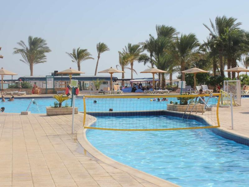 Outdoor pool with a volleyball net, sun umbrellas, and palm trees under clear sky.