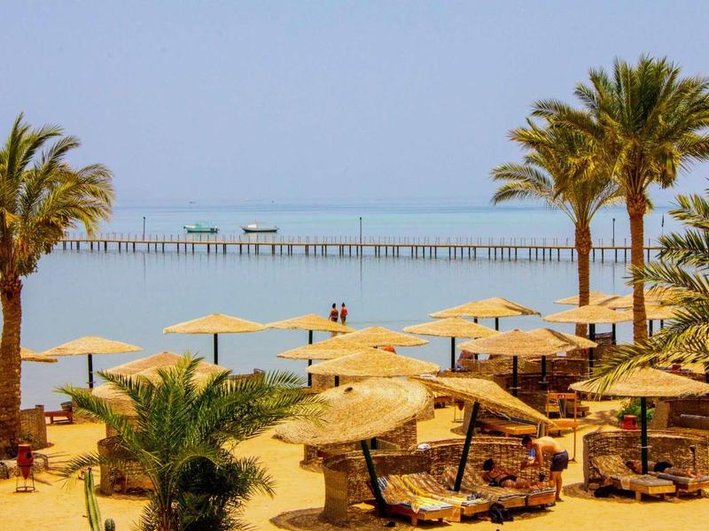 Beach resort with palm trees, sun umbrellas, lounge chairs, and a pier stretching into calm sea.