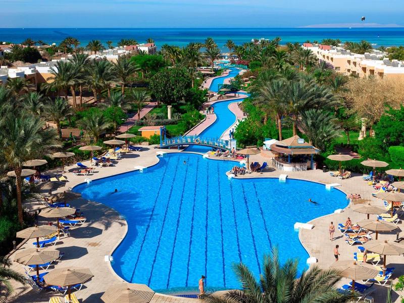 Large outdoor pool with winding waterway surrounded by palm trees and sun loungers at a beach resort.
