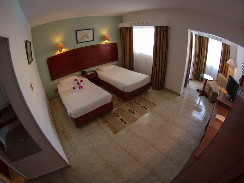 Hotel room with two single beds, beige tiled floor, wooden furniture, and large window with curtains.