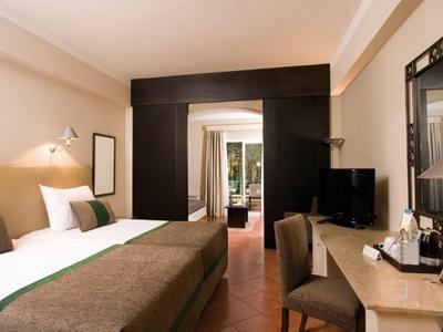 Hotelzimmer mit Doppelbett, Schreibtisch, Fernseher und offener Tür zu Balkon mit Grünblick.