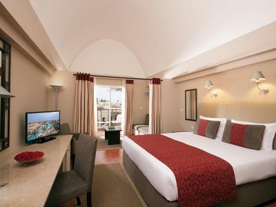 Hell eingerichtetes Hotelzimmer mit großem Bett, roten Akzenten und Blick auf Balkon.