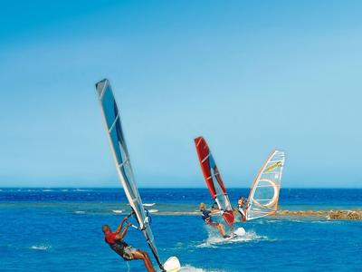 Drei Windsurfer fahren auf blauem Meer mit klar blauem Himmel und kleiner Insel im Hintergrund.