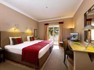 Helles Hotelzimmer mit Doppelbett, roten Akzenten, Schreibtisch, TV und Fenster mit Blick ins Grüne.