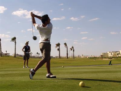 Golfspieler beim Abschlag auf grünem Rasen mit blauem Himmel und Palmen im Hintergrund.