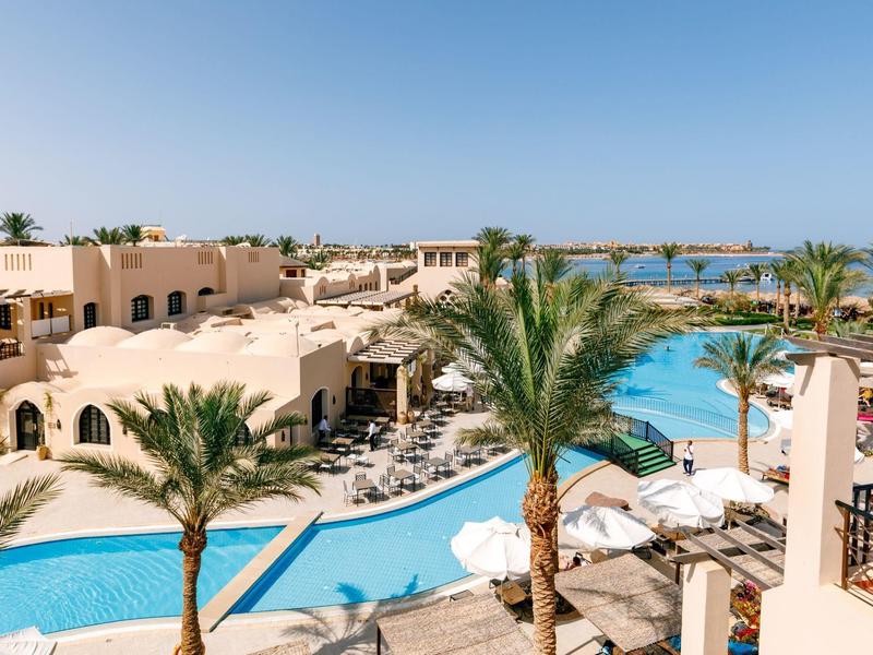 Large hotel pool with palm trees, sun loungers, and umbrellas under a clear blue sky.