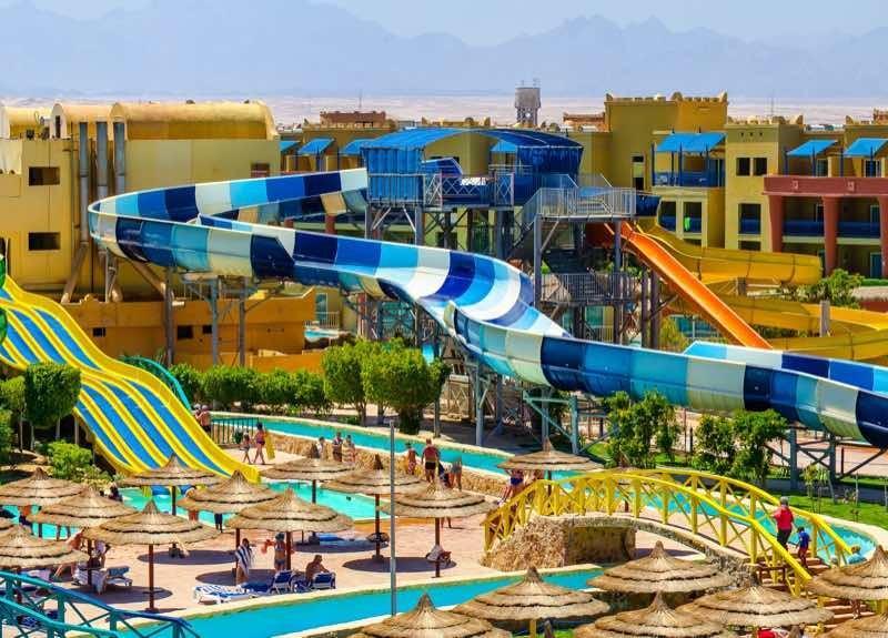 Grand parc aquatique avec toboggans colorés et palmiers devant des bâtiments d'hôtel.