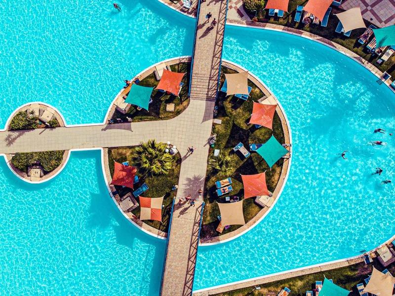 Grand piscine d'hôtel avec ponts et chaises longues, entourée de bâtiments et de palmiers.