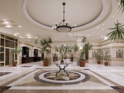 Ruime hotelhal met ronde plafond, planten en centrale tafel op marmeren vloer.