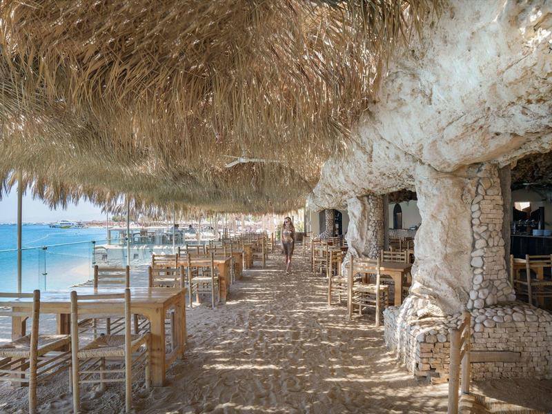 Restaurante de playa con mesas de madera bajo un techo natural de piedra junto al mar.