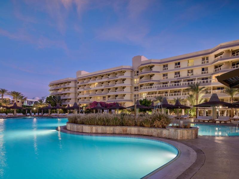 Hotel con piscina iluminada y área exterior al atardecer bajo cielo azul.