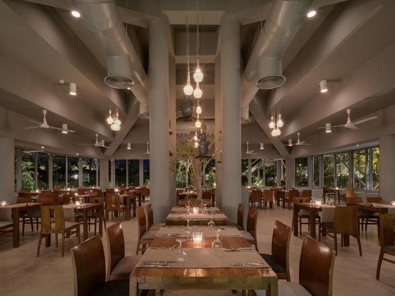 Gran comedor moderno con sillas y mesas de madera bajo conductos visibles y luz cálida.