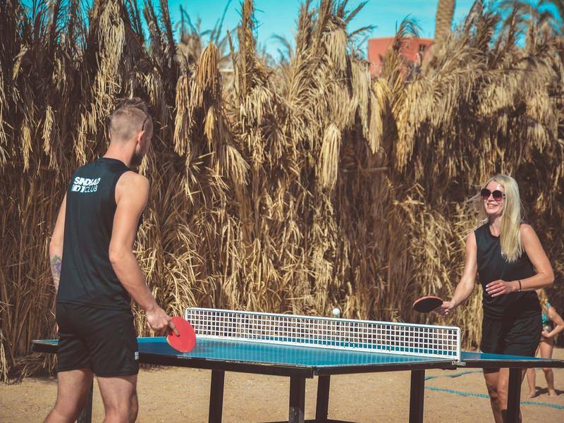 Dos personas juegan tenis de mesa al aire libre frente a una pared alta de cañas secas.