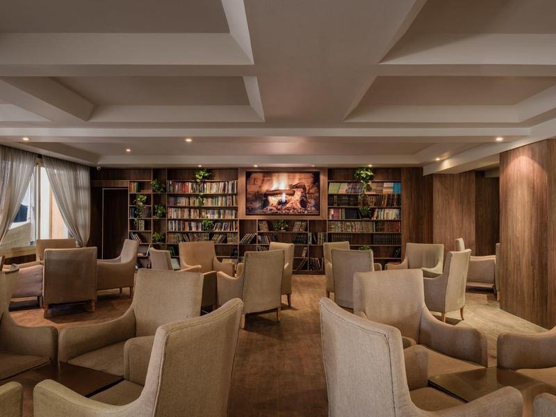 Cómoda sala de hotel con sillones beige, elementos de madera y una estantería con libros en la pared.