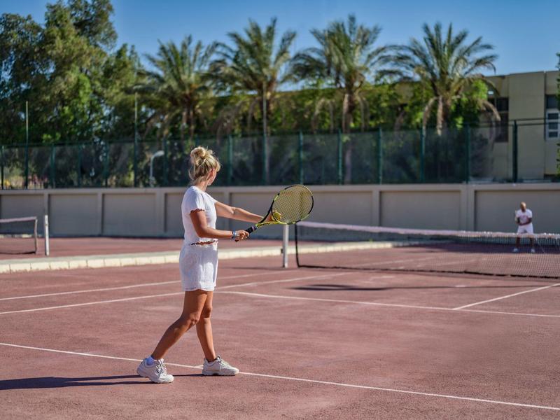 Dos personas juegan al tenis en una cancha al aire libre bajo un cielo despejado y palmeras de fondo.