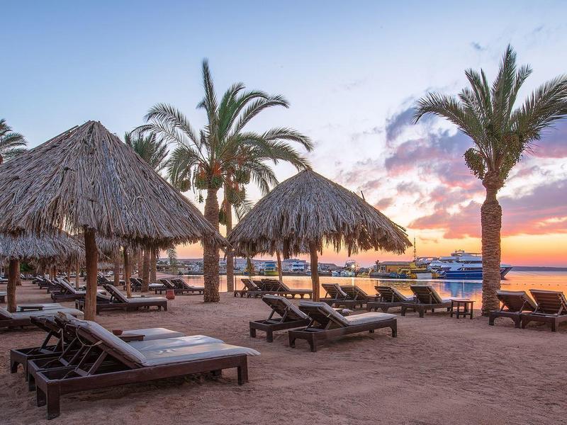 Atardecer en la playa con tumbonas bajo palmeras y techos de paja.
