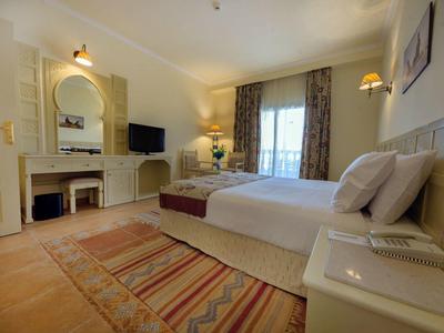 Helles Schlafzimmer mit Doppelbett, Teppichboden, Gardinen, Spiegel und Fernseher an der Wand.
