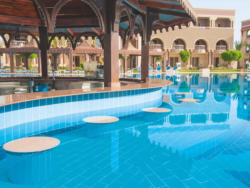 Modrý bazén s barovými stoličkami ve vodě vedle hotelu s oblouky.